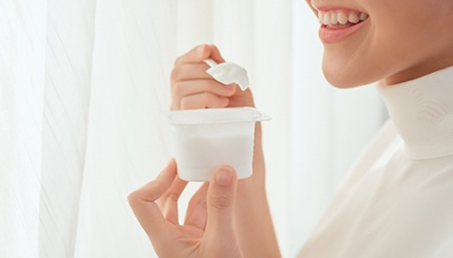 Kvinne smiler mens hun holder en yoghurt og spiser med skje."