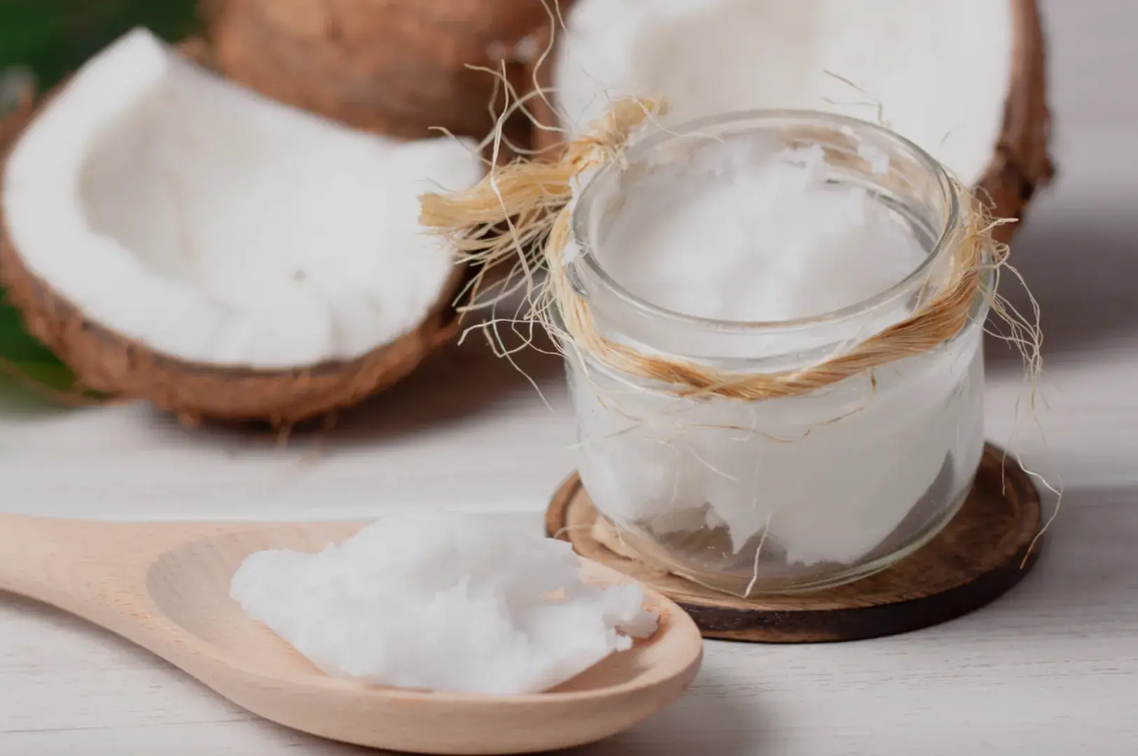 A small glass jar with coconut oil sits on a wooden surface, accompanied by a wooden spoon holding some of the oil, with coconut halves and green leaves in the background.