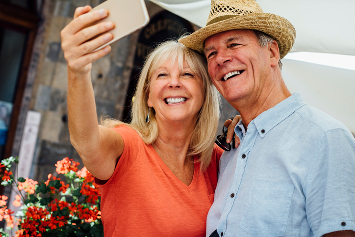 Blonde woman in an orange shirt snapping a selfie of herself and a man in a straw hat. 