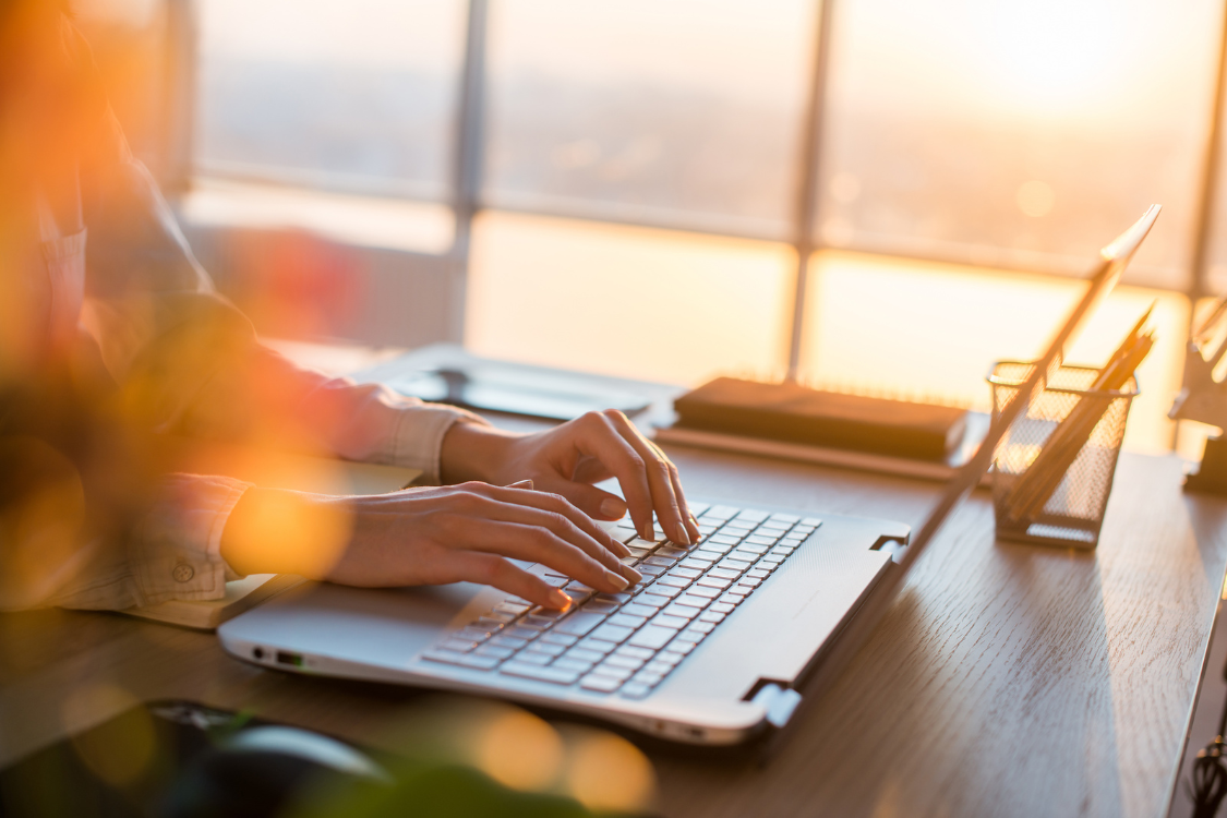 Woman typing on laptop at modern office desk during sunset, researching broker directories online