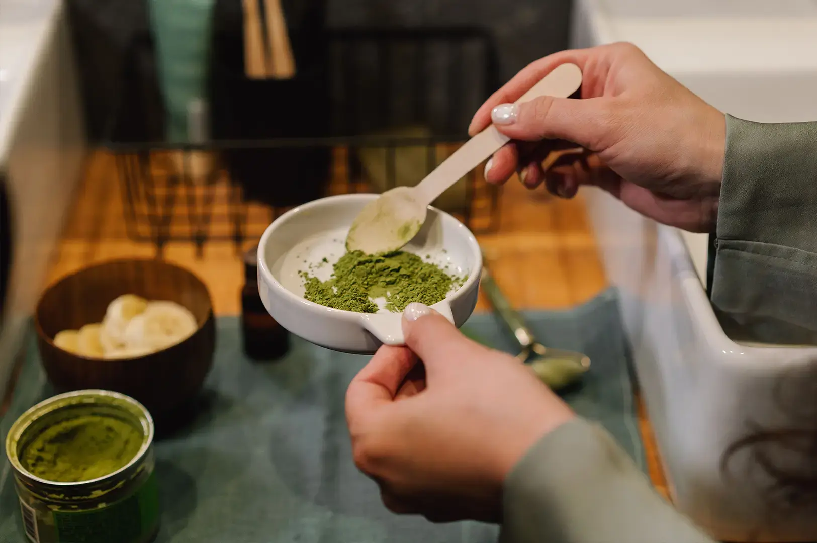 A person's hands are shown from a high angle, carefully using a light-colored wooden spoon to scoop or arrange fine, powdered green matcha into a small, round white dish, with a small wooden bowl of sliced fruit and a glass jar of matcha visible in the background.