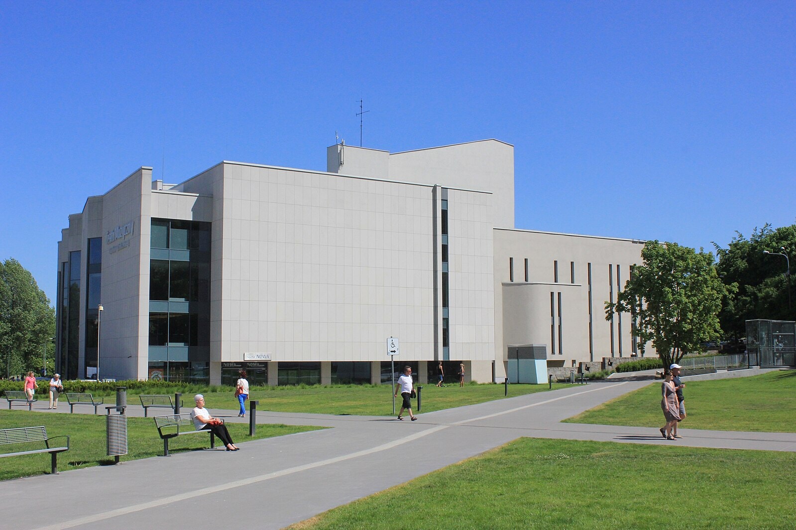 Budynek Teatru Muzycznego w Gdańsku. Szaro-beżowa konstrukcja otoczona skwerem, parkiem i zielenią. (źródło: https://commons.wikimedia.org/wiki/File:Gdynia_Teatr_Muzyczny.jpg)