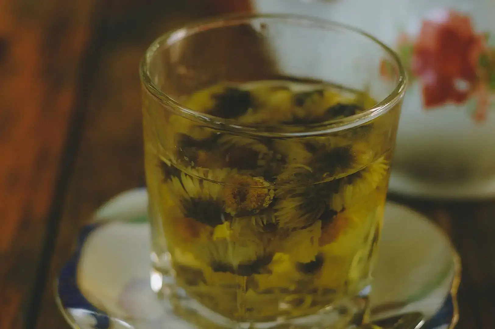 A warm, softly lit photograph of a small, straight-sided clear glass cup filled with light yellow tea, densely packed with floating chrysanthemum flower heads. The glass rests on a white ceramic saucer with blue and gold accents along the rim. The background is softly blurred, showing a dark wooden surface on the left and a glimpse of a white teapot decorated with red flowers on the right.