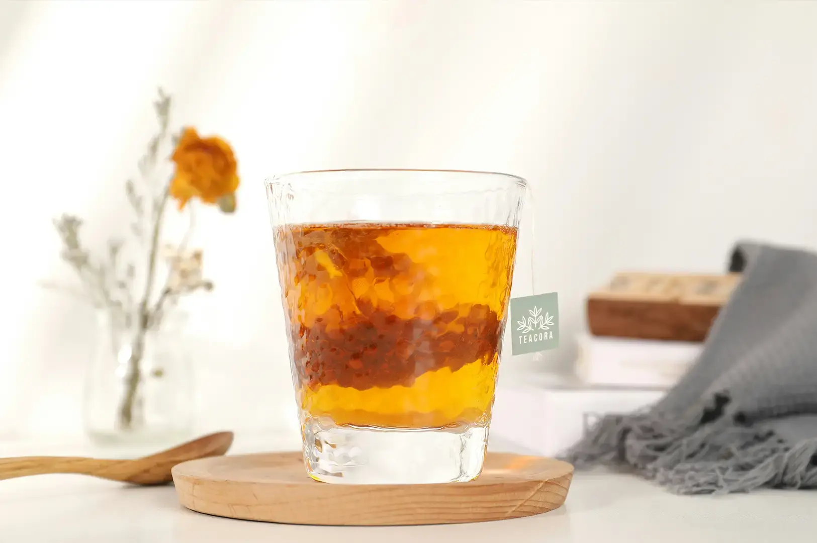 A clear, textured glass tumbler filled with bright amber-colored tea sits on a round wooden coaster. A tea bag tag with a green leaf logo and the word "TEACORA" hangs from the side of the glass. In the soft-focus background, a small glass vase holding a dried orange flower and delicate stalks sits to the left, while a gray fringed cloth and a stack of books rest on the right side of the clean white surface.
