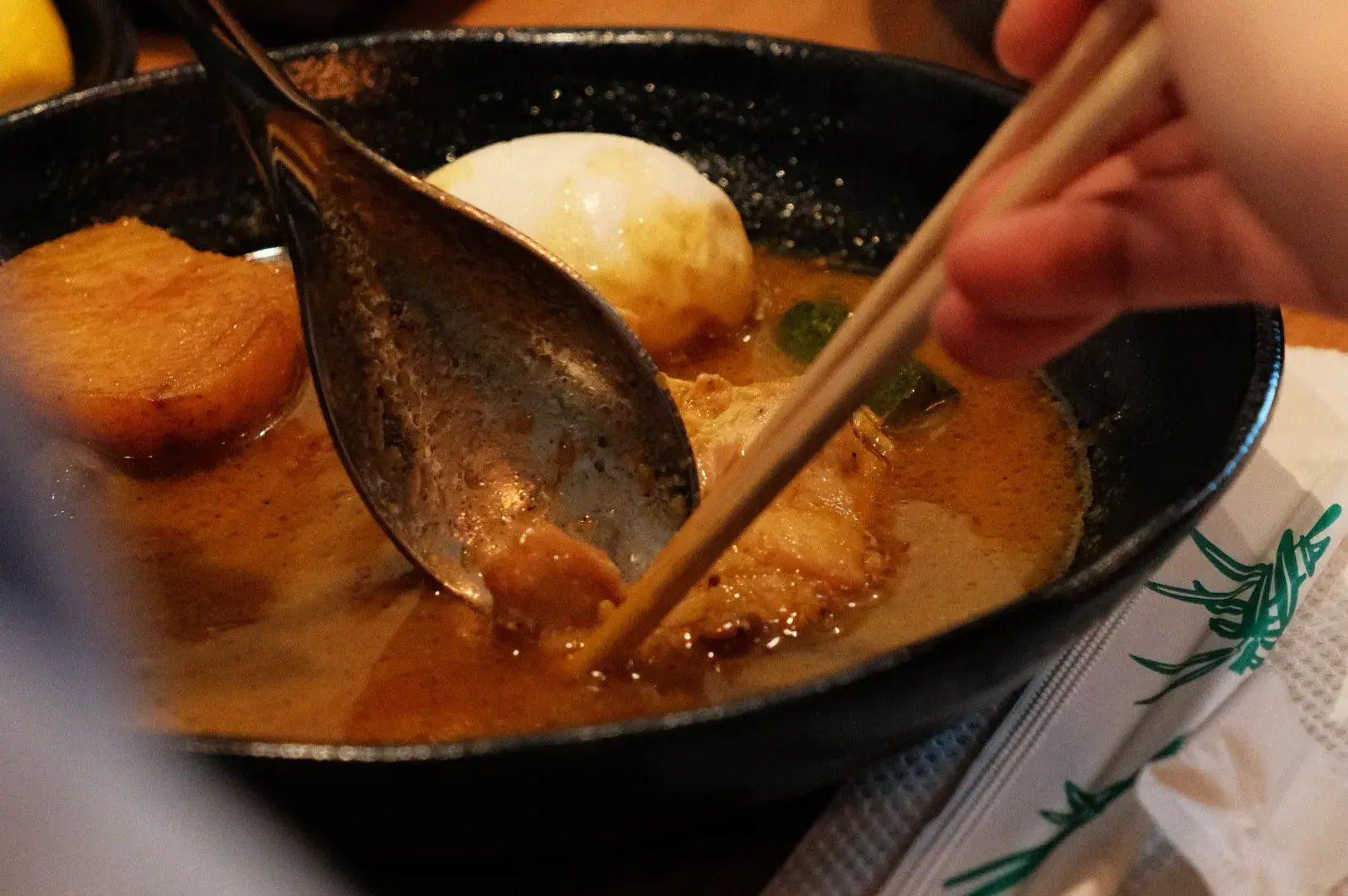 A close-up of a bowl of ramen with slices of pork, a soft-boiled egg, and someone using chopsticks to scoop food.