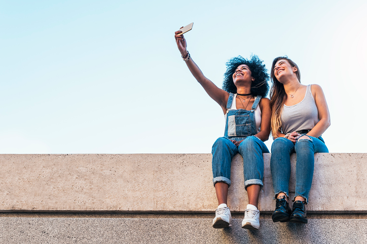 Two happy young women sitting on a cement wall snapping a selfie.