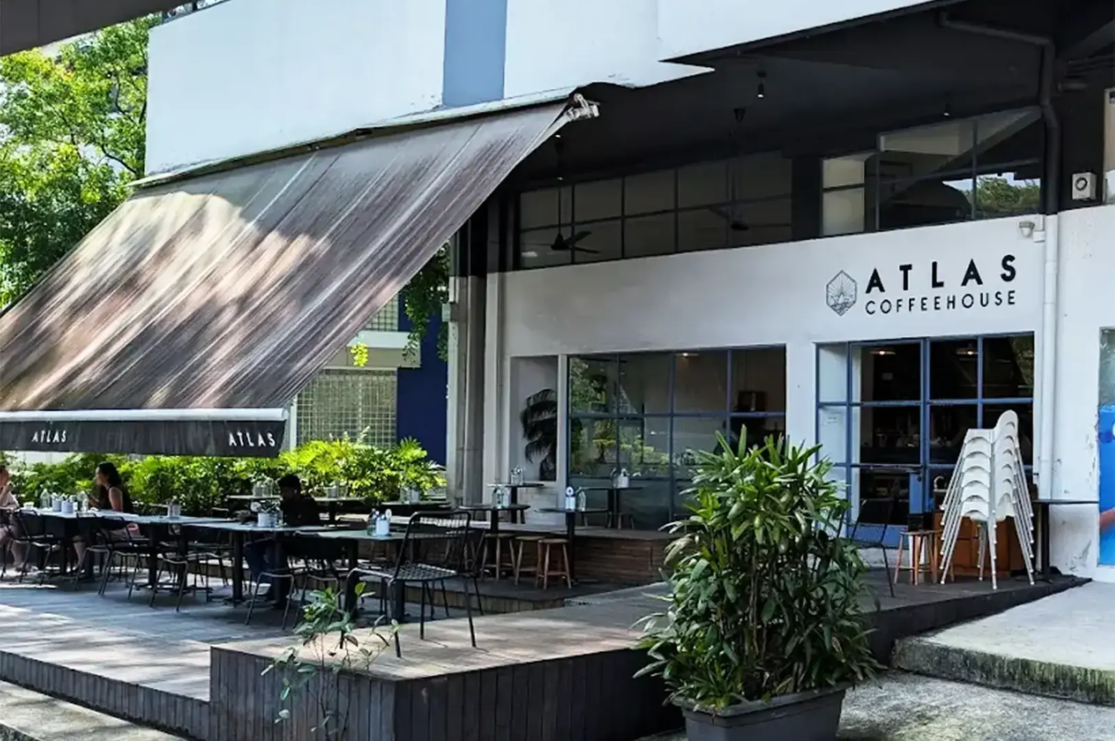 Outdoor view of Atlas Coffeehouse featuring a shaded patio with tables and chairs. The facade is modern, surrounded by greenery, creating a relaxed ambiance.