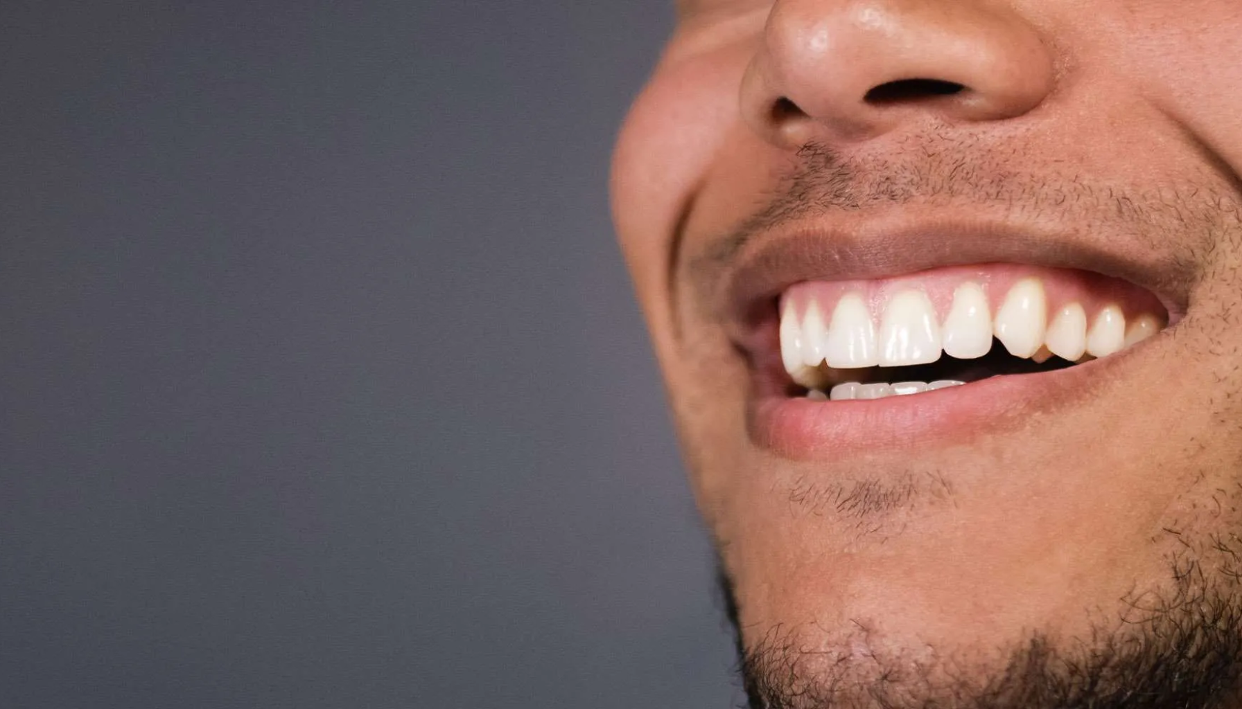 Tannkjøttet Trekker Seg Tilbake: Smil Med Lengre Tenner? 10 Nærbilde av smilende mann med hvite, friske tenner og kort skjegg mot grå bakgrunn. tannkjøtt trekker seg tilbake