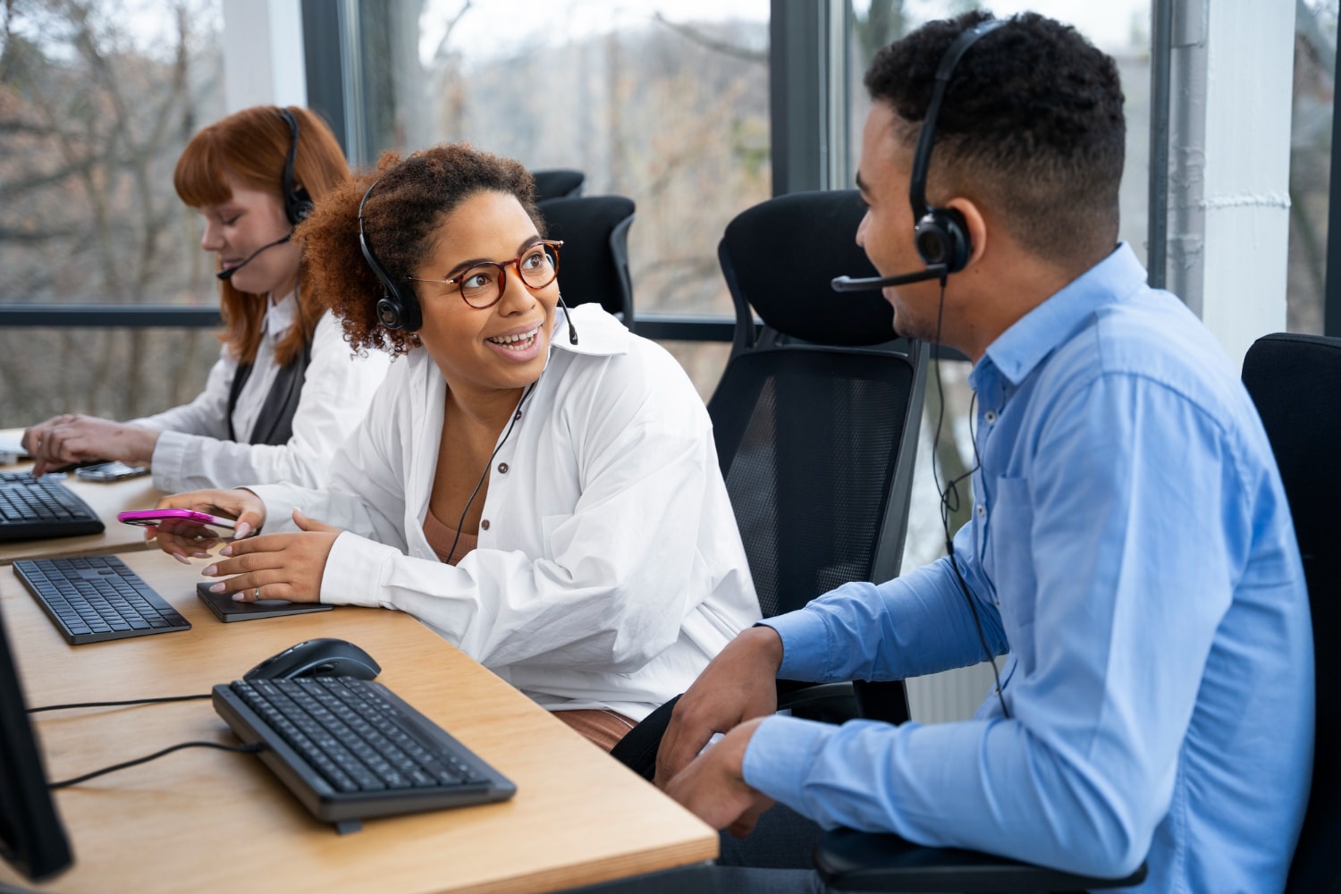 A group of agents wearing headsets in a sleek, well-lit office interact enthusiastically. One woman gestures while speaking to a colleague, demonstrating teamwork powered by scalable contact center platforms.