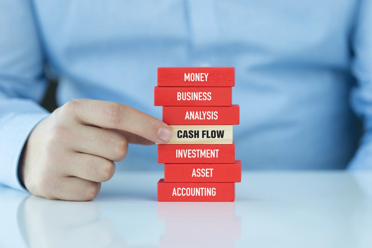 A hand points to a stack of blocks with financial terms. The blocks read: Money, Business, Analysis, Cash Flow, Investment, Asset, and Accounting.