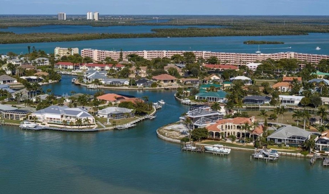 The image captures a stunning panoramic sunset view from a luxury home in Factory Bay, showcasing a private dock with boats gently bobbing on the water, framed by the beautiful hues of the sunset. This serene scene highlights the idyllic lifestyle available in Marco Island, FL, offering breathtaking views and direct access to the bay.