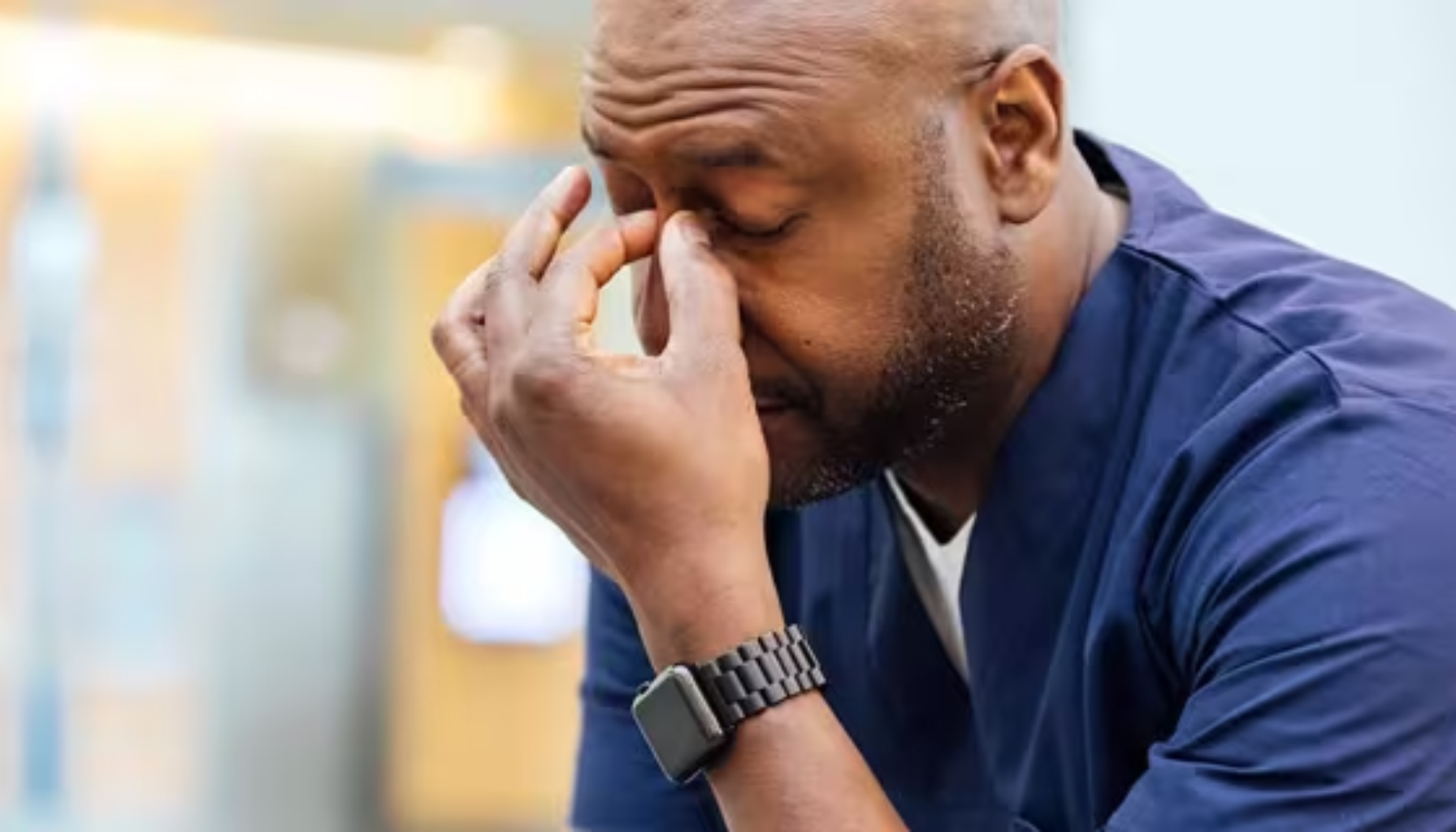 Spenningshodepine: Kan Komme Fra Kjeven 11 Mann i helseuniform sitter bøyd fremover og klyper neseroten med hånden, tydelig sliten eller stresset på jobb innendørs