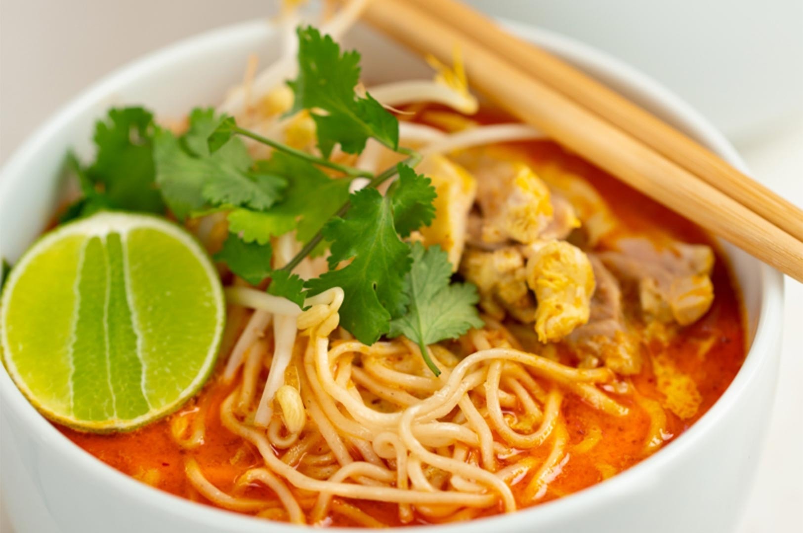 A bowl of spicy coconut curry noodle soup (Laksa) served with thin rice noodles, bean sprouts, cilantro, and a lime wedge.