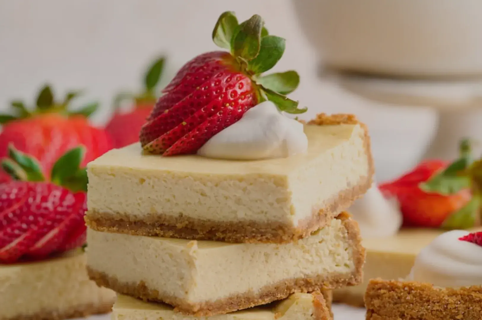 A stack of creamy cheesecake squares topped with a fresh strawberry and a dollop of whipped cream, surrounded by additional strawberries.