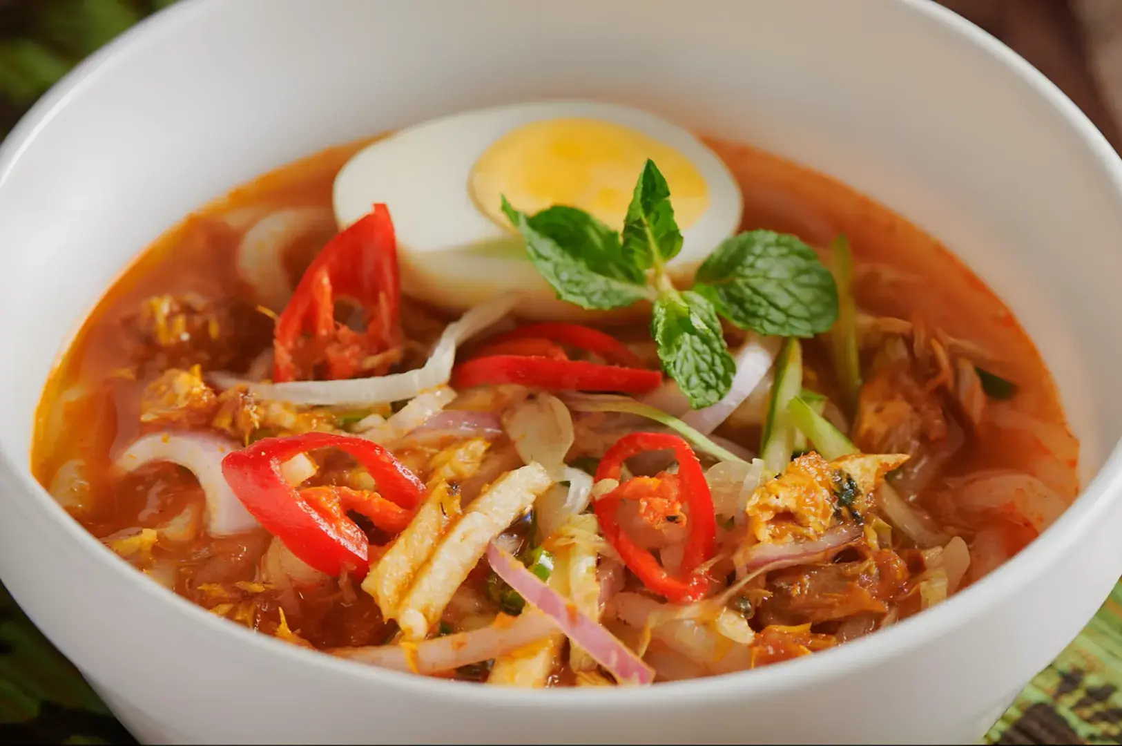 A bowl of spicy soup with noodles, shredded chicken, vegetables, sliced red chili, mint leaves, and a halved boiled egg.