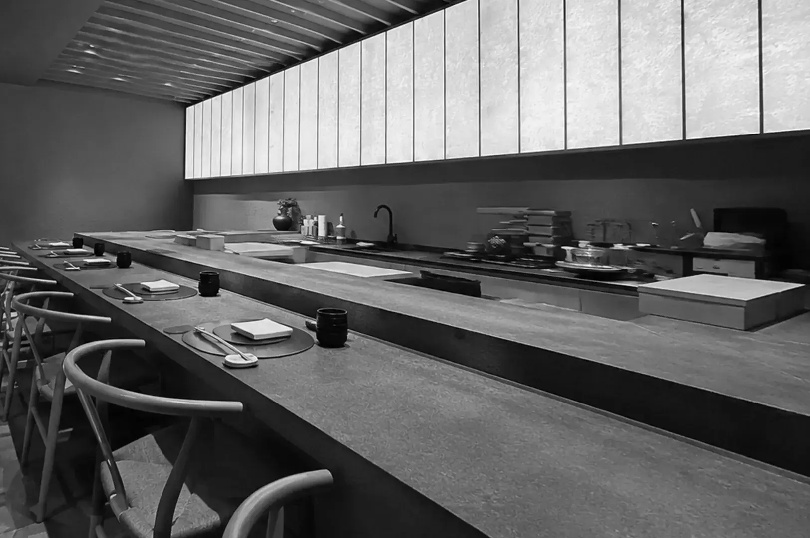 Minimalist sushi restaurant with a long counter, open preparation area, individual place settings, and a large illuminated window.