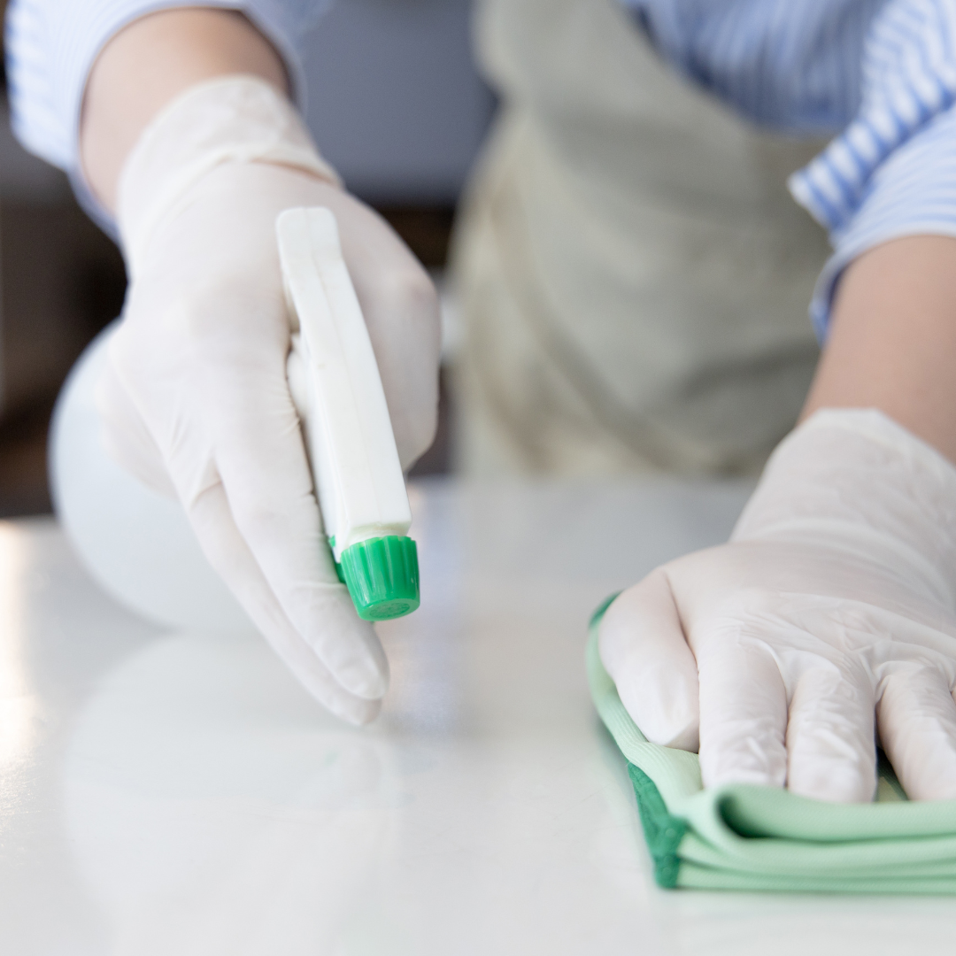 person with an apron, gloves, spray bottle, and damp cloth wiping down a table