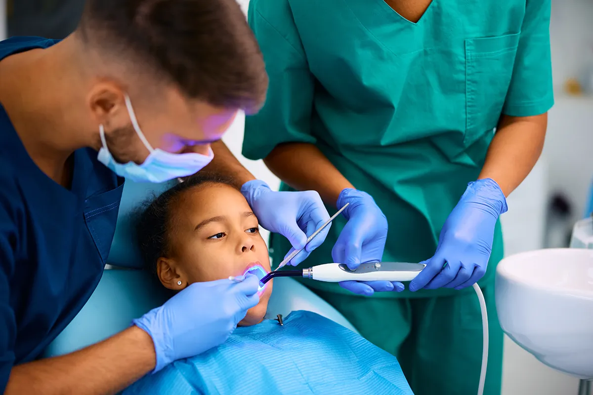 Young child receiving dental care from two professionals using examination tool.