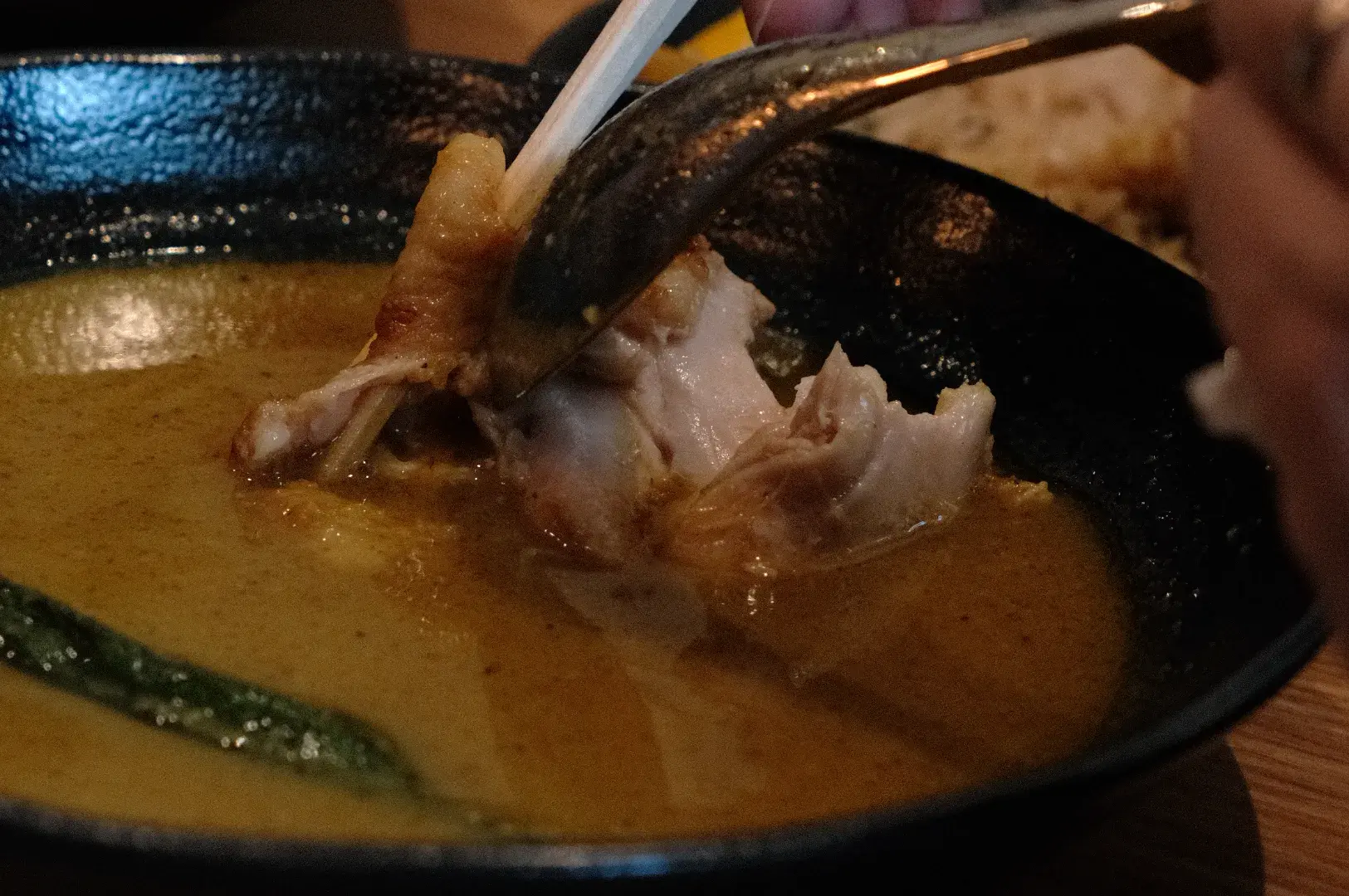 A close-up of a hand using chopsticks to pick up a piece of chickenfrom a bowl of soup curry.