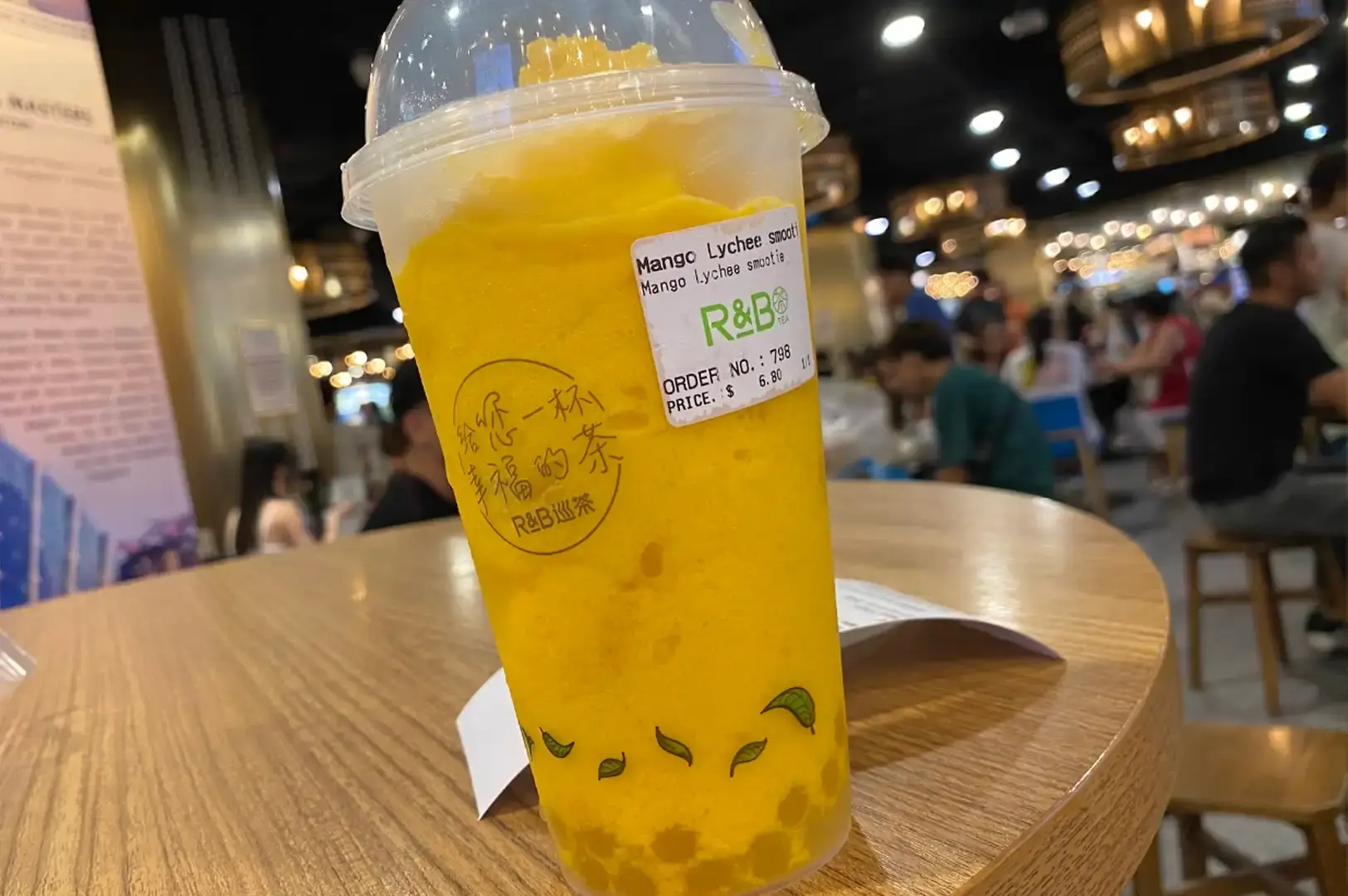 A close-up shot of a tall clear plastic cup filled with a bright yellow Mango Lychee Smoothie from R&B Tea, topped with a domed lid and featuring golden pearls settled at the bottom, resting on a light wooden table inside a bustling food hall.