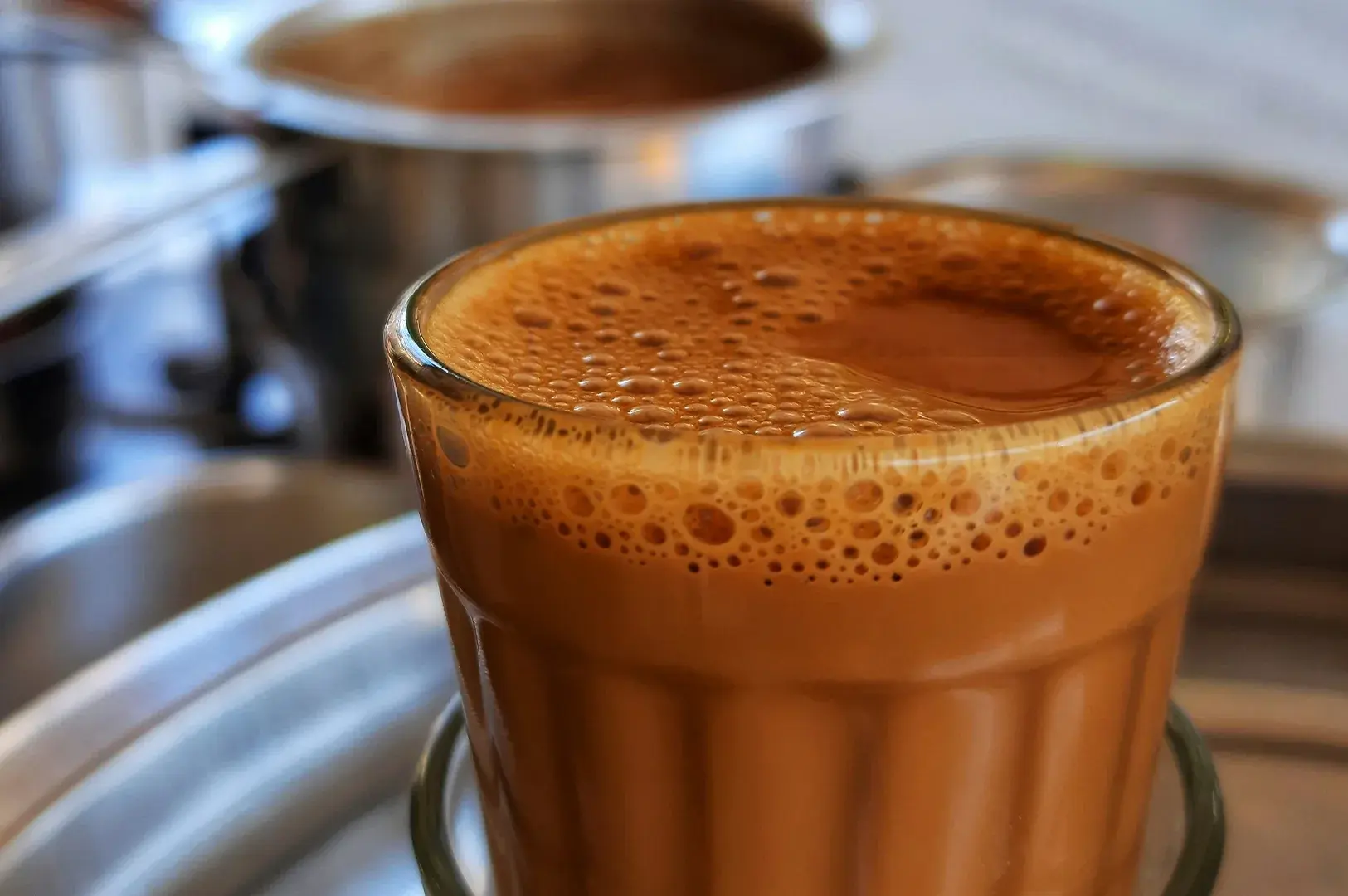 A shallow depth-of-field close-up of a small, vertically ribbed glass filled to the brim with rich, frothy, golden-brown masala chai, with a blurred metal pot visible in the background, accentuating the texture and warmth of the drink.