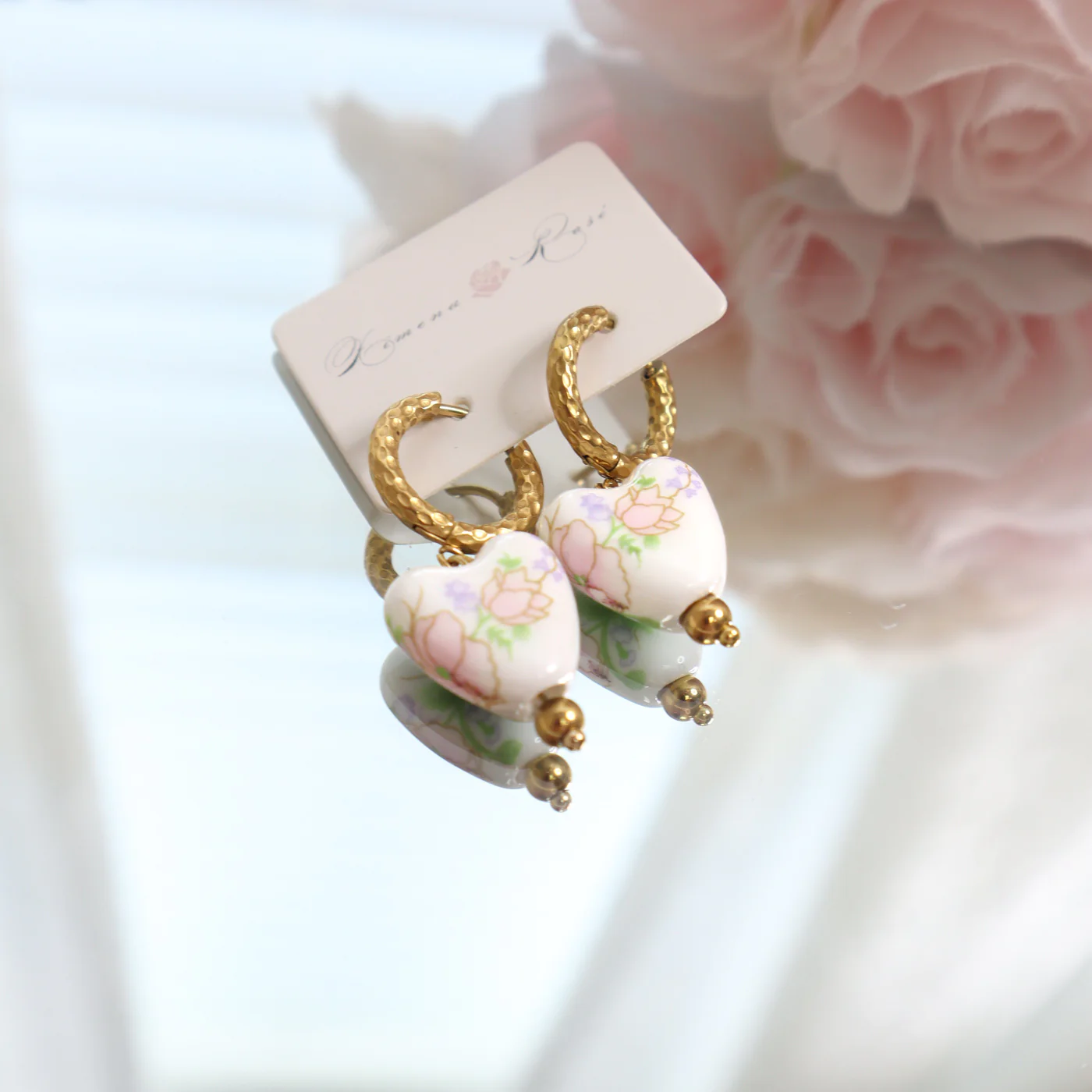 Small hoop earrings with a floral white heart pendant laid on a mirror, accompanied by pink roses at the top right corner and a 'Ximena Rose' brand tag.