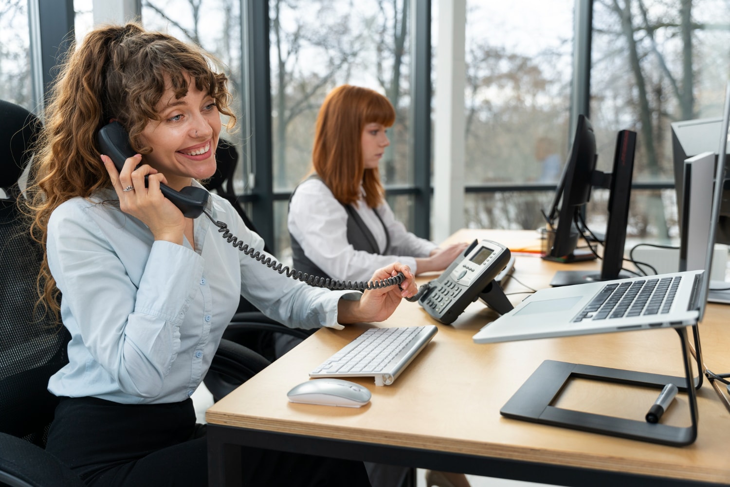 A smiling customer service representative in a modern office using a corded phone while working on a computer. Another colleague is visible in the background. This illustrates Business phone solutions and Hosted PBX Solutions enhancing customer engagement.