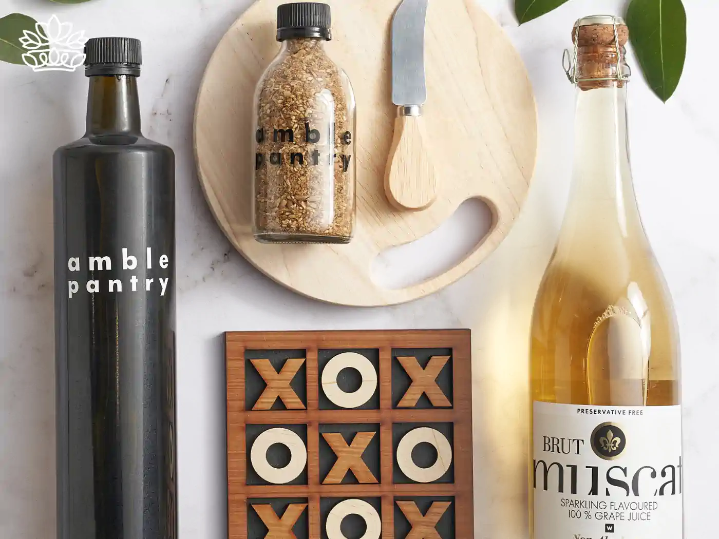 A carefully curated gift set featuring pantry items like olive oil and spices, a bottle of grape juice, a wooden cheese board, a knife, and a tic-tac-toe game, arranged beautifully on a marble surface. Fabulous Flowers and Gifts