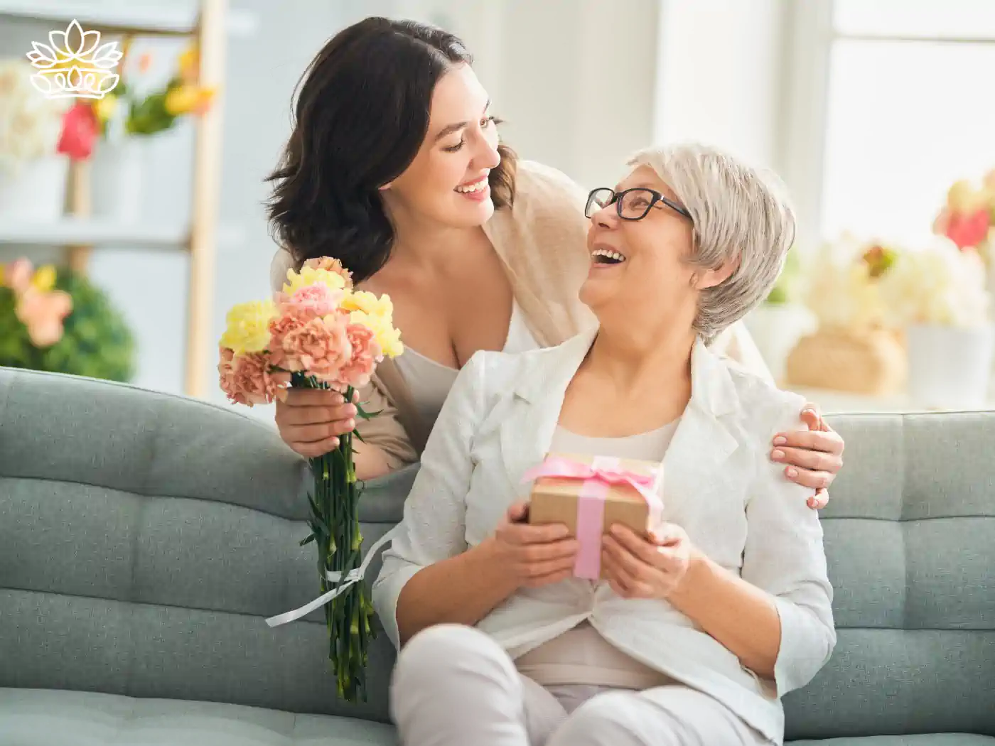 A daughter presenting her elderly mother with a bouquet and a gift while they sit on a sofa. Fabulous Flowers and Gifts. Mother's Day Flowers.