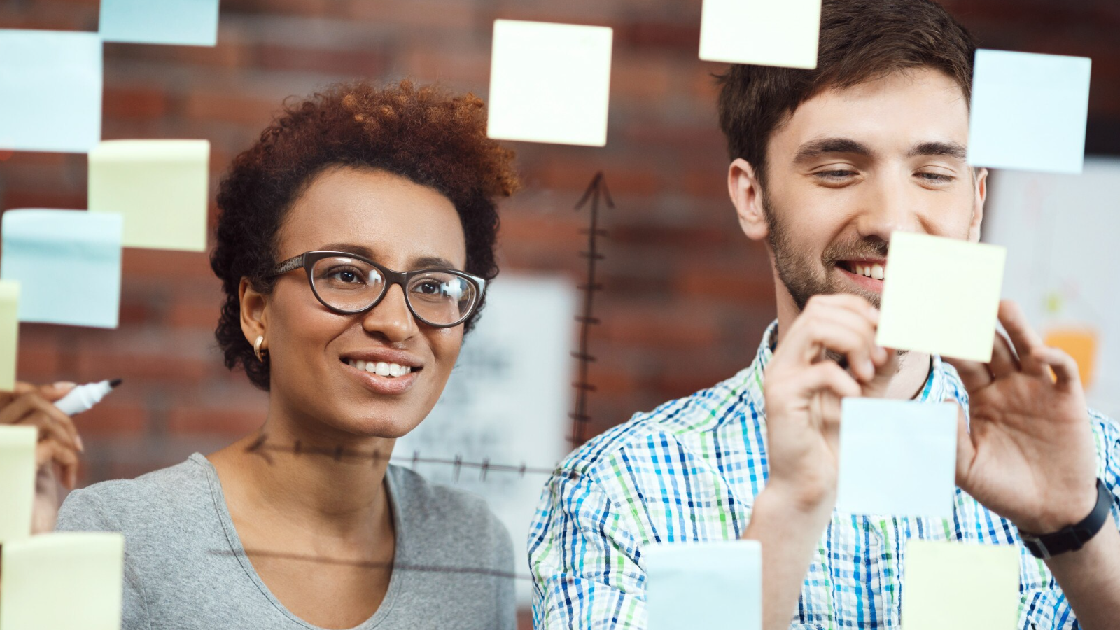 Two coworkers adding sticky notes to a glass board, visualizing how teams collaborate and align on communication goals.