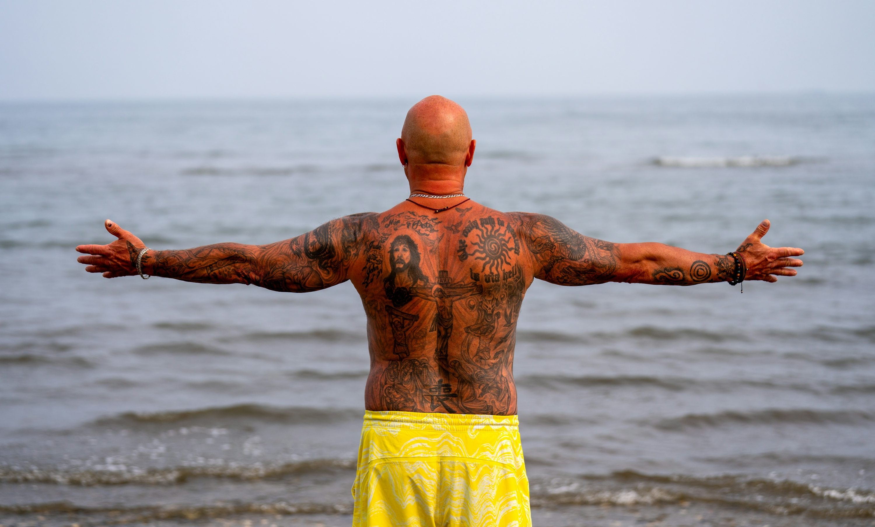 a man with large tattooed area on his back standing in front of the ocean. 