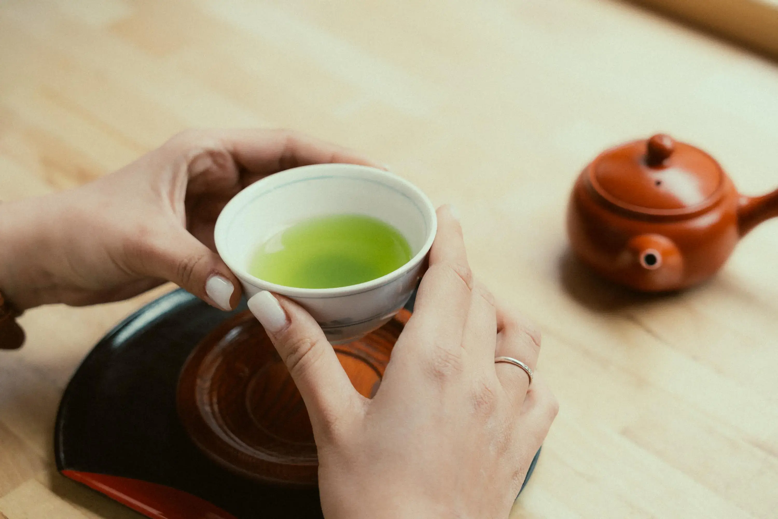A person with manicured nails holds a small white ceramic cup filled with vibrant green tea above a wooden saucer. In the blurred background, a small, reddish-brown clay teapot rests on the light wooden table.