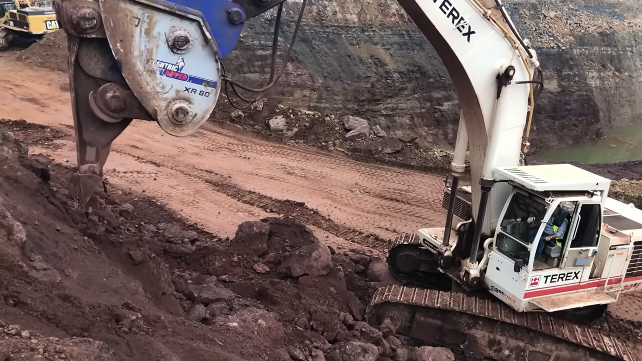 A large blue and white Terex excavator with a hydraulic hammer attachment digging into the ground at a construction site