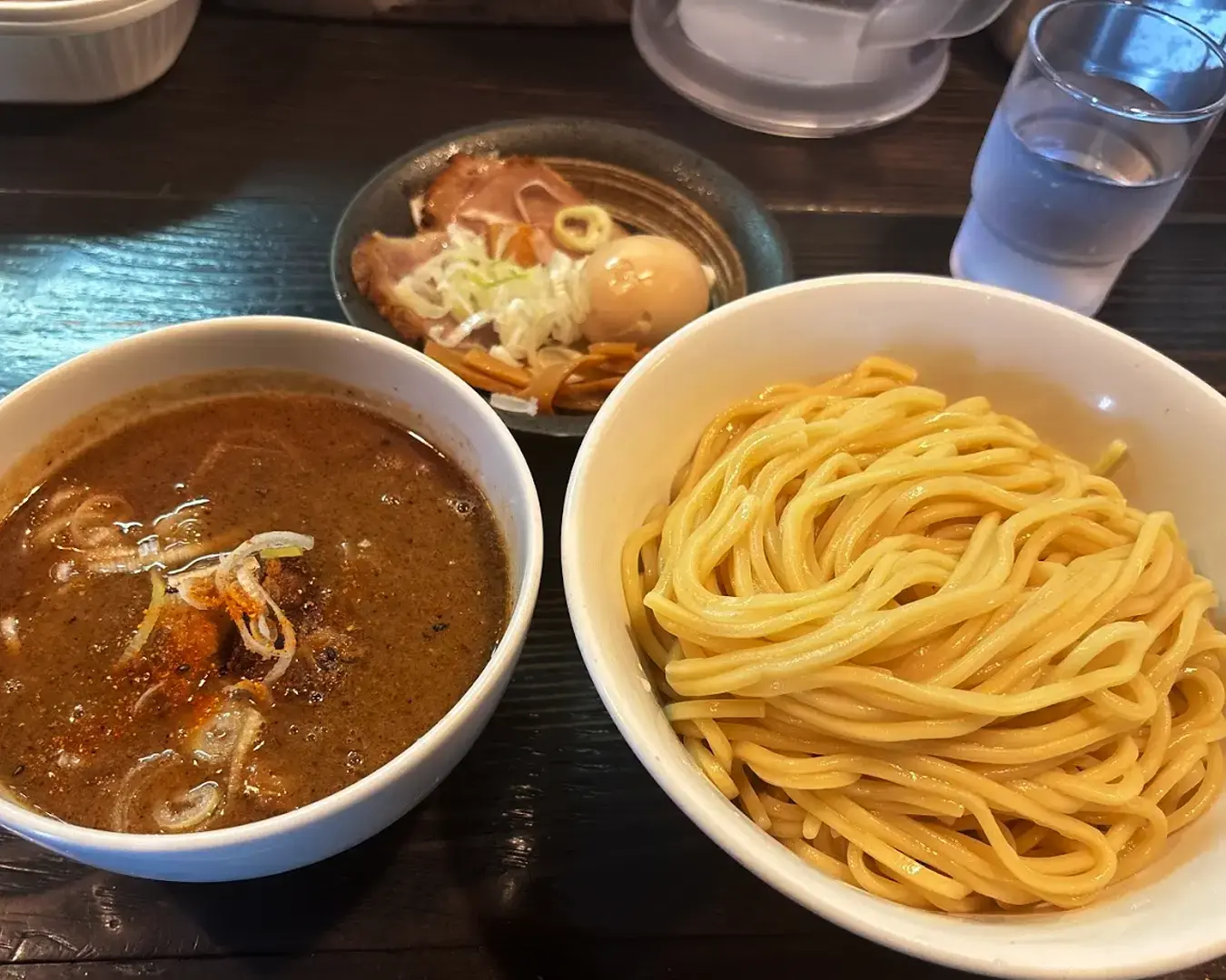 A bowl of thick noodles sits next to a bowl of rich, brown dipping sauce topped with green onions. In the background, there is a dish with sliced pork, boiled egg, and vegetables. A water glass is placed nearby on the dark wooden table, creating a warm, inviting dining scene.