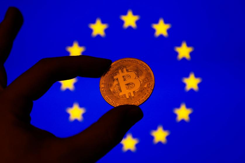A image of a hand holding a Bitcoin with the European Union flag behind it, suggesting that Bitcoin can help the Euro. 