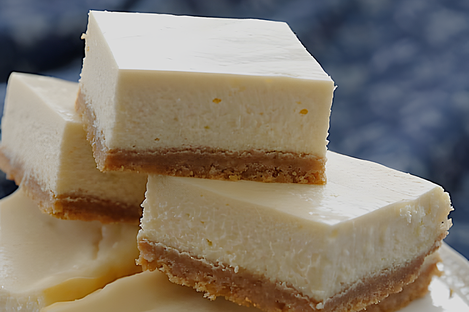 Stack of creamy cheesecake squares with a golden brown crust, neatly arranged. The background is a soft, out-of-focus dark blue cloth.