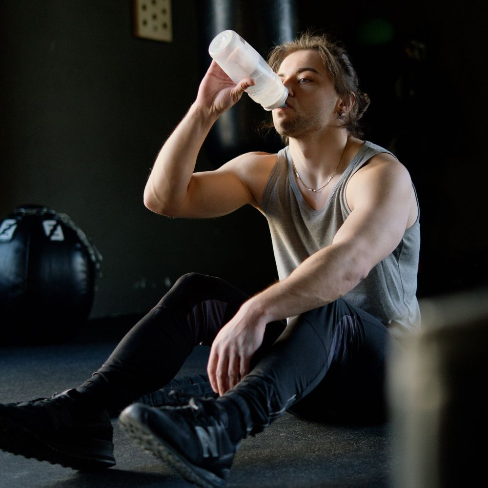 A person drinking a glass of water with a bottle of Total War Pre-Workout supplement in the background