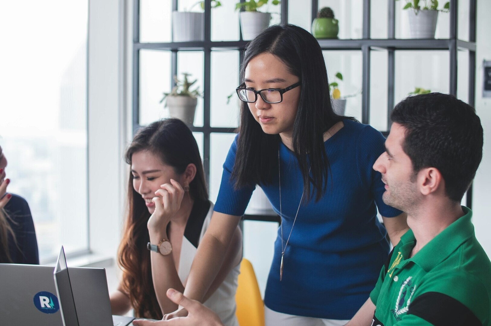 A woman in glasses stands, pointing at a laptop screen, engaging with two colleagues seated and smiling. Background includes plants on shelves, creating a collaborative and vibrant setting.