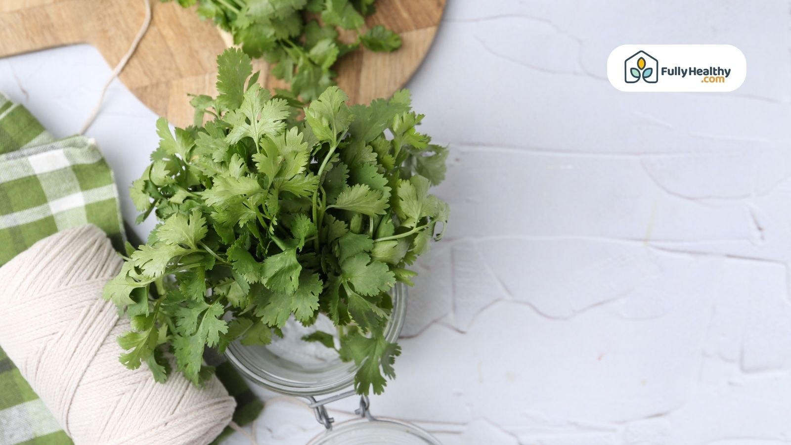 Fresh green herbs in a glass jar on a white surface beside a green checkered cloth and a roll of string