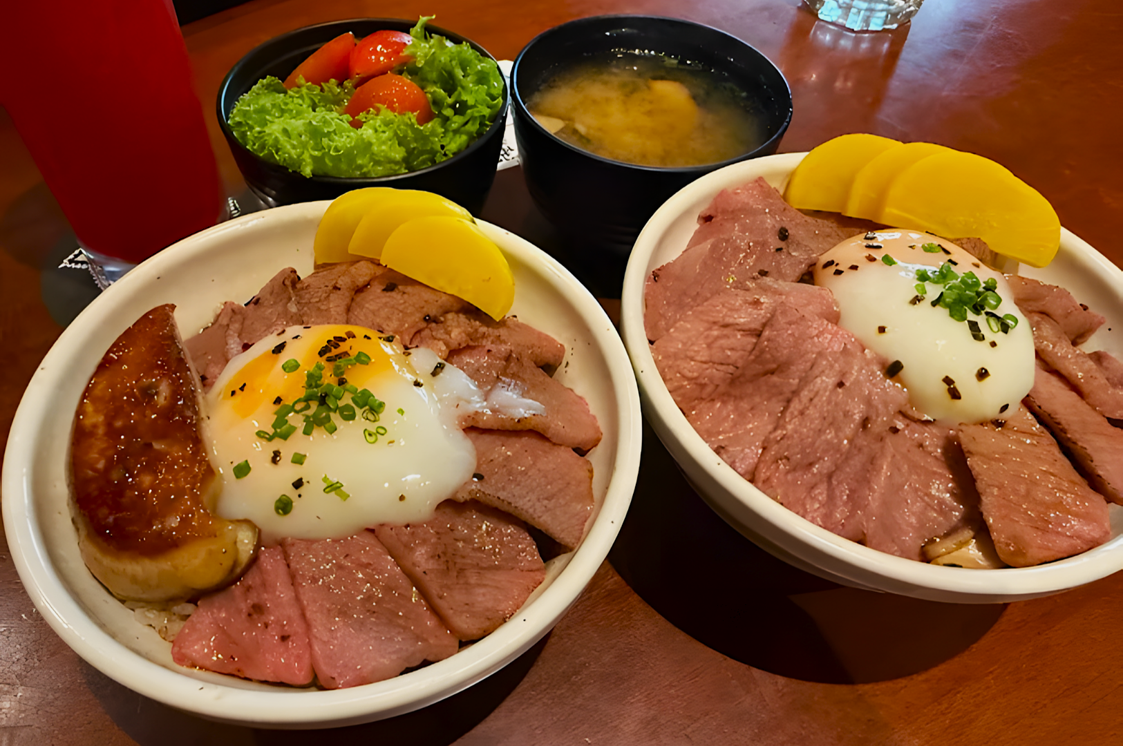 Two bowls of beef donburi topped with poached eggs and pickles are on a table alongside a fresh green salad, miso soup, and a red drink.