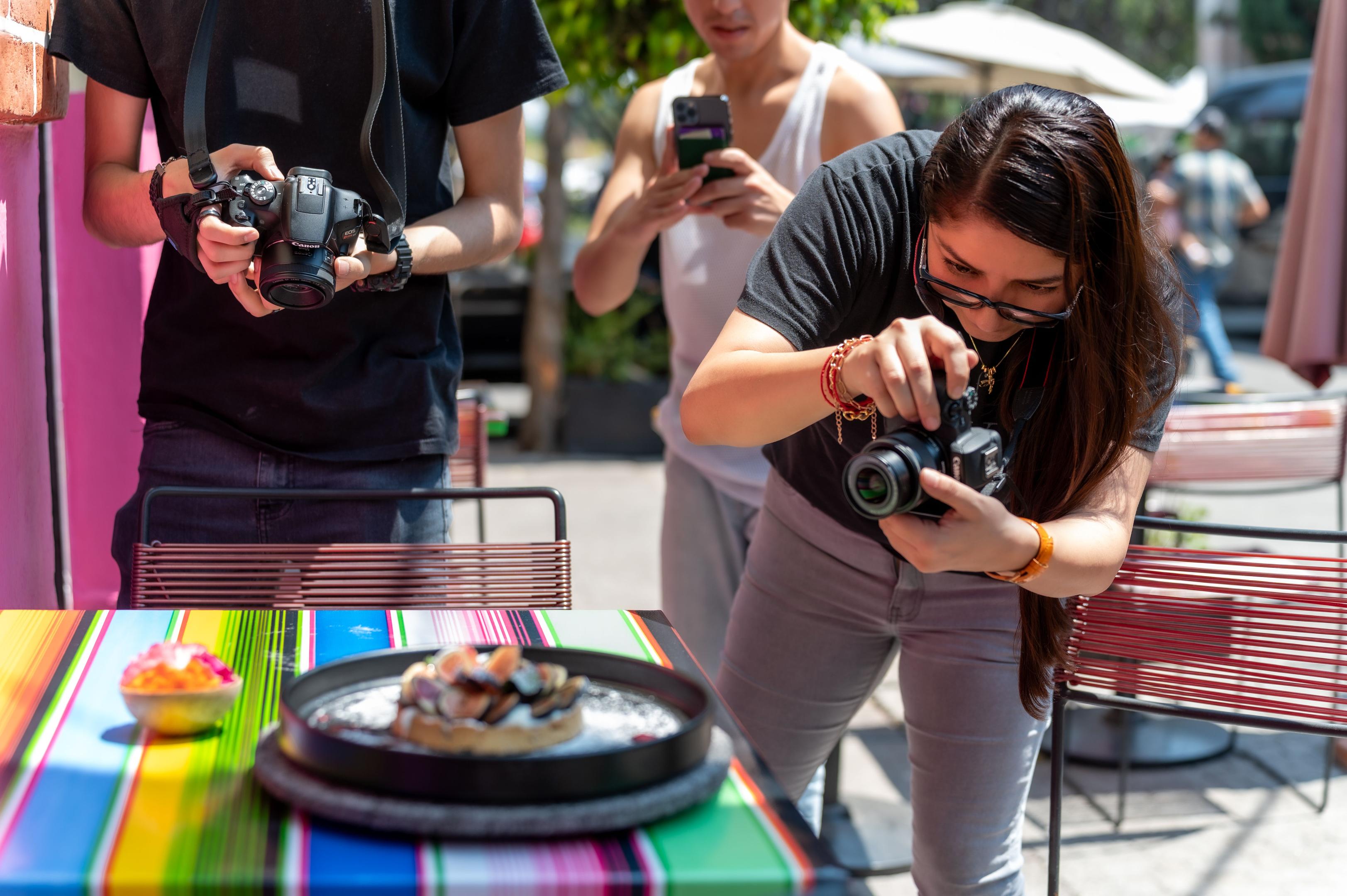 A group of restaurant diners is gathered around a table, excitedly taking a photo of their beautifully plated menu items with a smartphone, showcasing their food for social media platforms. This moment captures the essence of user-generated content, as guests engage in sharing their dining experience, contributing to the restaurant's social media presence.