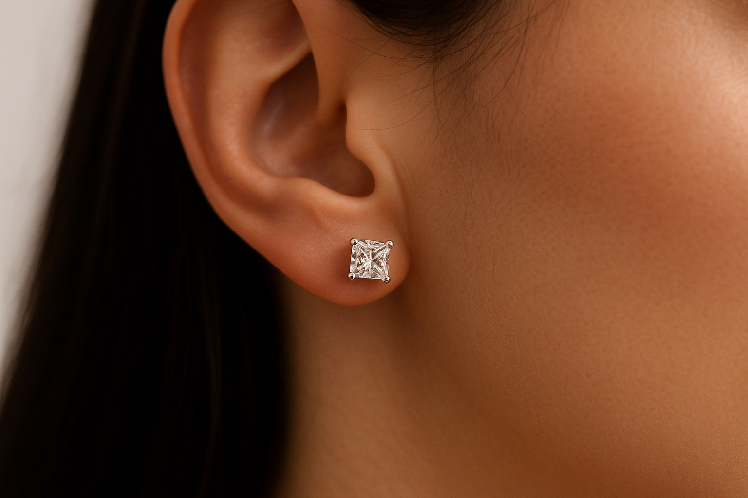 Artimas vaizdas į stilingos merginos ausį, puoštą balto aukso auskaru su tobulai simetrišku Princess formos deimantu, ryškiais kvadratiniais kraštais ir spindesiu.
