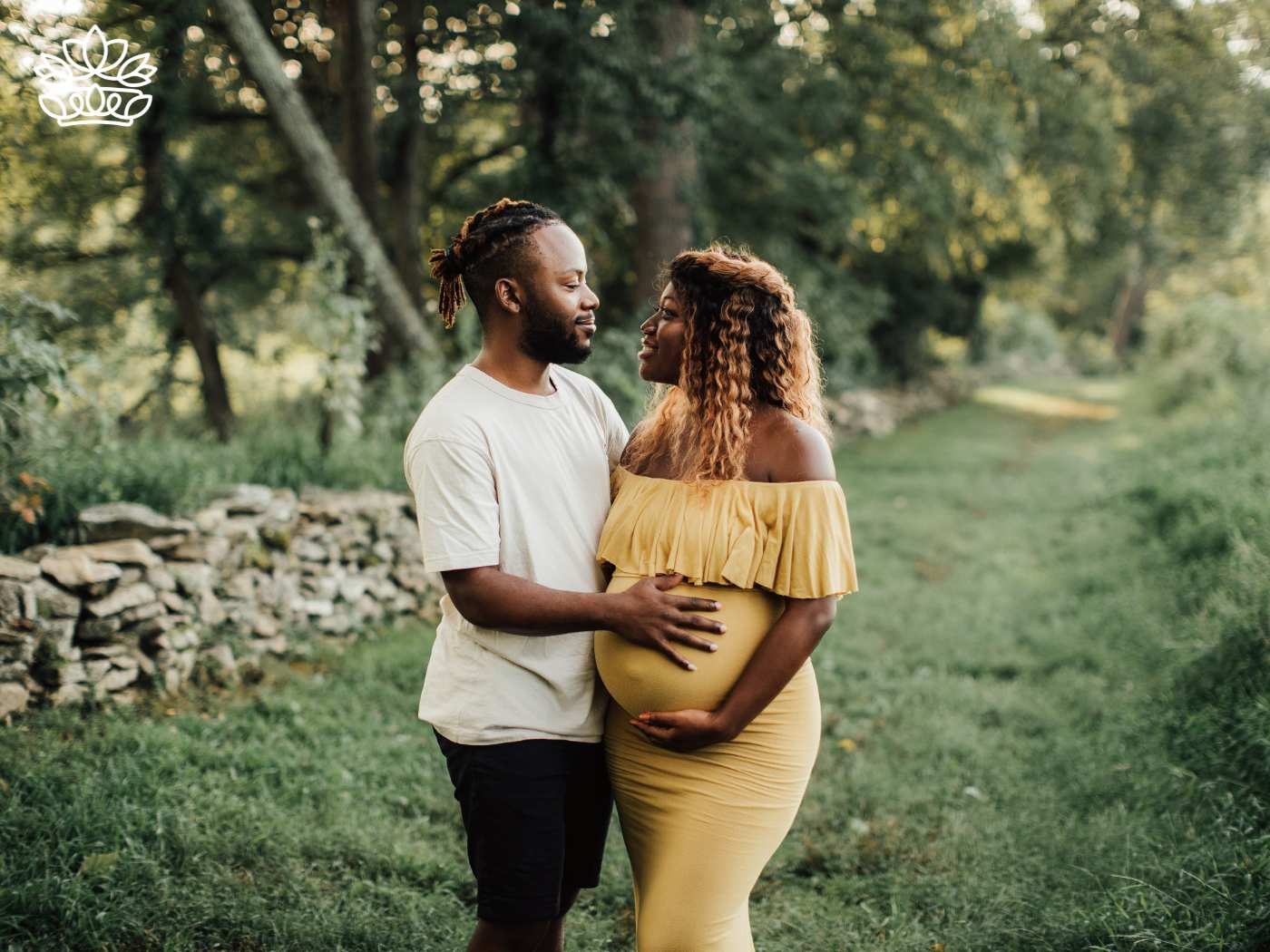 A tender moment captured in a serene outdoor setting, with a man gently holding his pregnant partner, who is radiant in a yellow dress. They share a loving gaze, surrounded by the natural beauty of a lush green landscape. A heartwarming scene for celebrating new beginnings with Fabulous Flowers and Gifts. Gift Boxes for wife. Delivered with Heart.