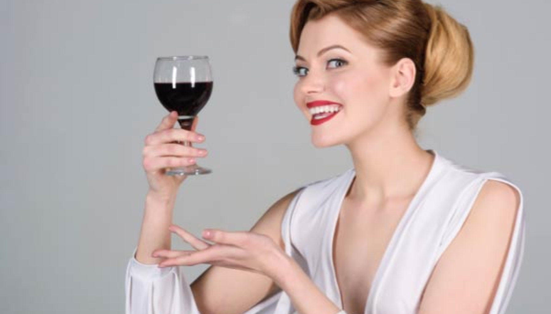 Kvinne i hvit kjole smiler bredt og holder opp et glass rødvin med entusiasme.