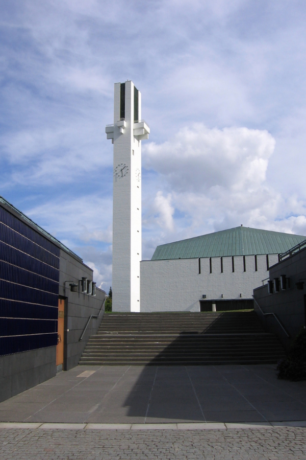 Exterior view of a modernist building with white brick upper walls, long horizontal windows framed in wood, and a dark vertical-tile facade section in the foreground.
