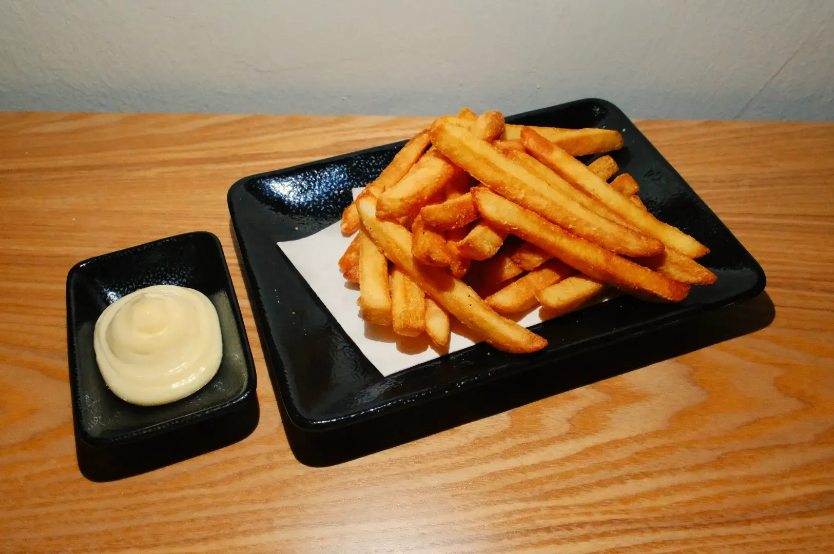 A plate of golden-brown French fries next to a small bowl of mayonnaise on a wooden table.