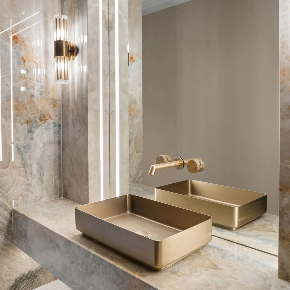 Luxurious bathroom vanity area featuring a brushed gold rectangular vessel sink, wall-mounted gold faucet, and marble-effect surfaces with integrated lighting.