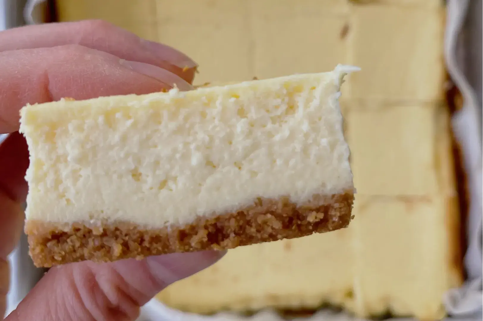 A close-up of a hand holding a slice of cheesecake with a graham cracker crust. The cheesecake appears smooth and creamy, with a light yellow color.