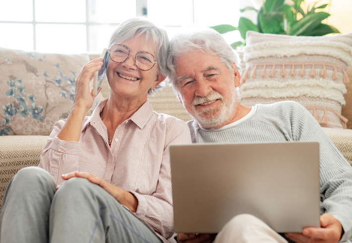 Mature couple sitting on the floor using a laptop and talking on a cell phone. 