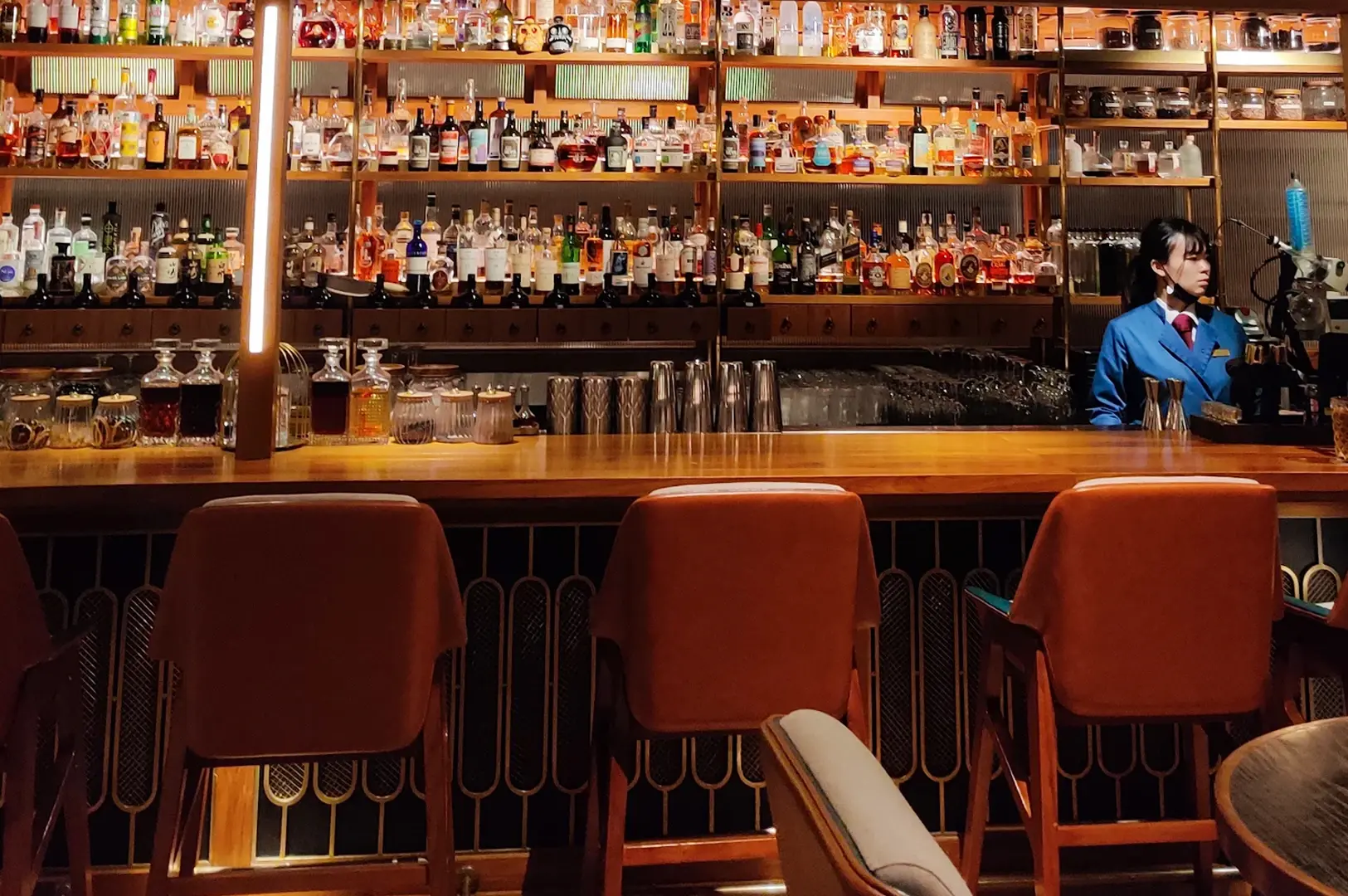 Eye‑level straight‑on shot of a stylish cocktail bar interior featuring a long wooden counter, upholstered bar stools, and shelves lined with assorted liquor bottles, with a bartender working behind the bar.